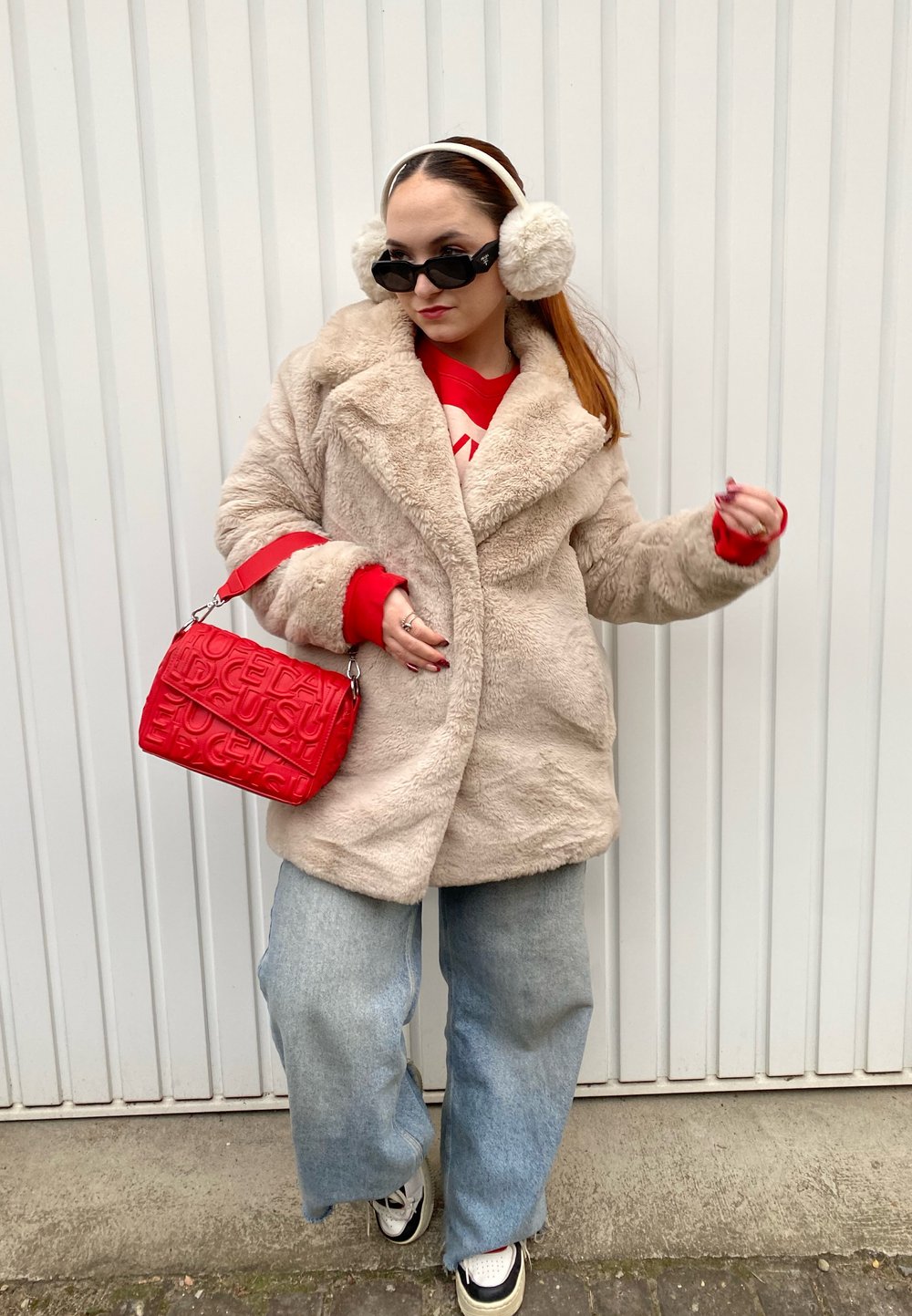 Μπεζ ψεύτικο γούνινο παλτό με μεγάλο γιακά, κόκκινο πουλόβερ από κάτω, μπλε παντελόνι φαρδύ, μαύρα γυαλιά ηλίου, κόκκινη τσάντα και λευκά αθλητικά παπούτσια.