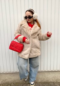 Μπεζ ψεύτικο γούνινο παλτό με μεγάλο γιακά, κόκκινο πουλόβερ από κάτω, μπλε παντελόνι φαρδύ, μαύρα γυαλιά ηλίου, κόκκινη τσάντα και λευκά αθλητικά παπούτσια.