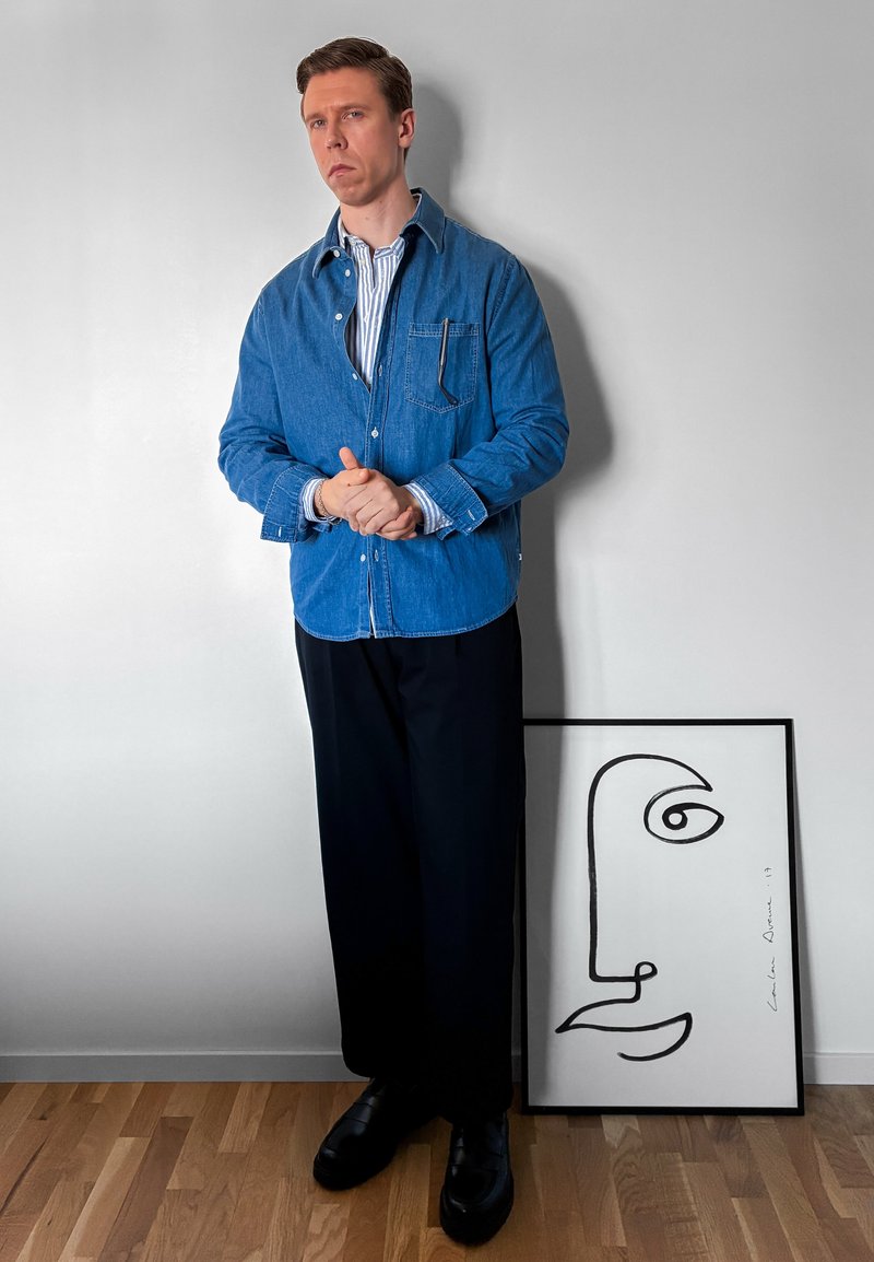 Homme portant une chemise en denim bleu et un pantalon noir se tient près d'une œuvre d'art minimaliste en noir et blanc représentant un visage abstrait appuyée contre le mur.