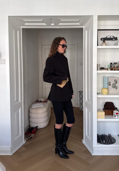 Mujer con blazer negro oversize, pantalones cortos de ciclismo negros, botas negras hasta la rodilla y gafas de sol en una habitación moderna con estanterías y decoración.