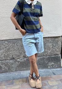 Persona vistiendo una camisa a rayas azul y verde con cuello blanco, pantalones cortos de mezclilla claros, pantuflas mullidas con correas negras, de pie contra una pared texturizada.