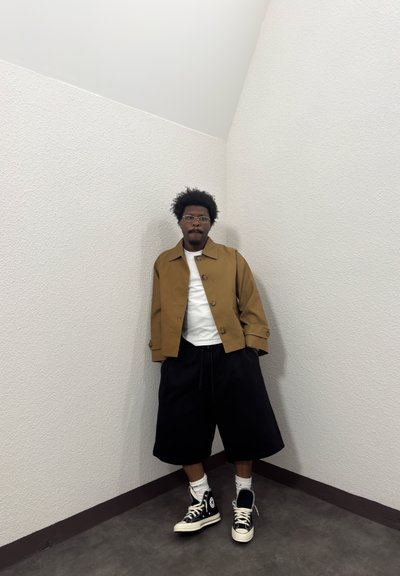 Hombre con gafas que lleva una chaqueta marrón, camisa blanca, pantalones cortos negros y anchos, y zapatillas negras de caña alta, de pie contra una pared blanca texturizada en una esquina.