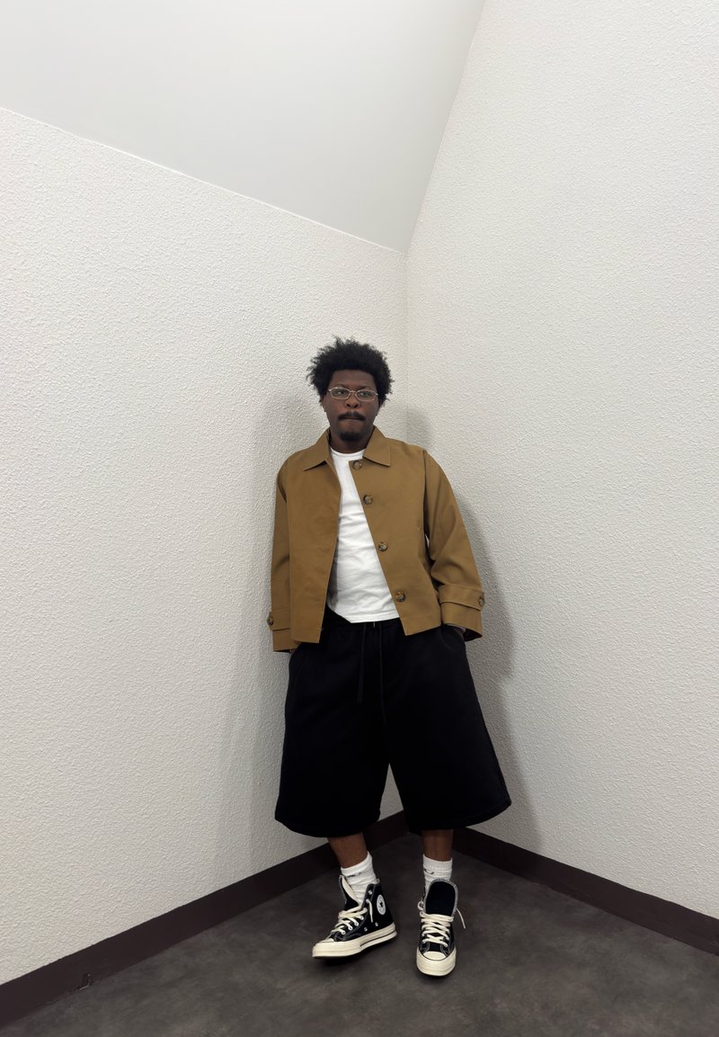 Mann med briller iført en brun jakke, hvit skjorte, svarte vide shorts og svarte høye sneakers som står mot en strukturert hvit hjørnevegg.