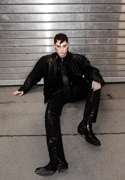 Chaqueta de cuero negra sobre una camisa negra y corbata, combinada con pantalones negros ajustados y botas de tobillo brillantes. Textura suave, acentos de hardware mínimos.