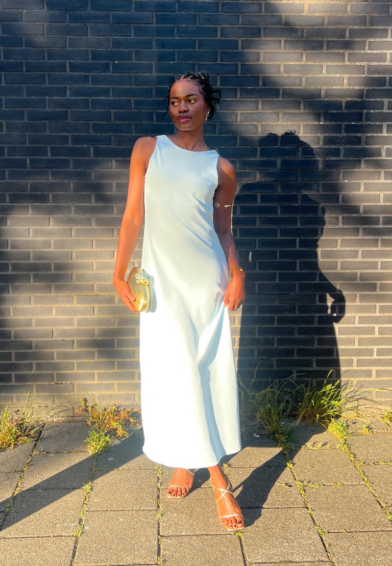 Világoskék, ujjatlan maxi ruha sima textúrával. A viselője egy arany színű clutch táskát tart a kezében. A szettet egy pántos, nude szandál egészíti ki.