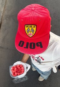 Rdeča kapa z logotipom, ki vsebuje šahovske zastave in napis "AL RACING"; drži plastično posodo, napolnjeno s koščki rožnatega lubenice.
