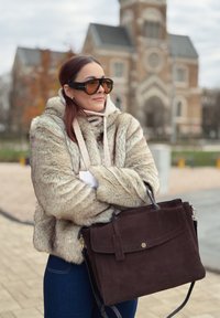 Braune Wildlederhandtasche mit elegantem Design, einem Tragegriff oben und verstellbarem Schulterriemen. Das Modell trägt einen flauschigen beigen Hoodie und eine Sonnenbrille.