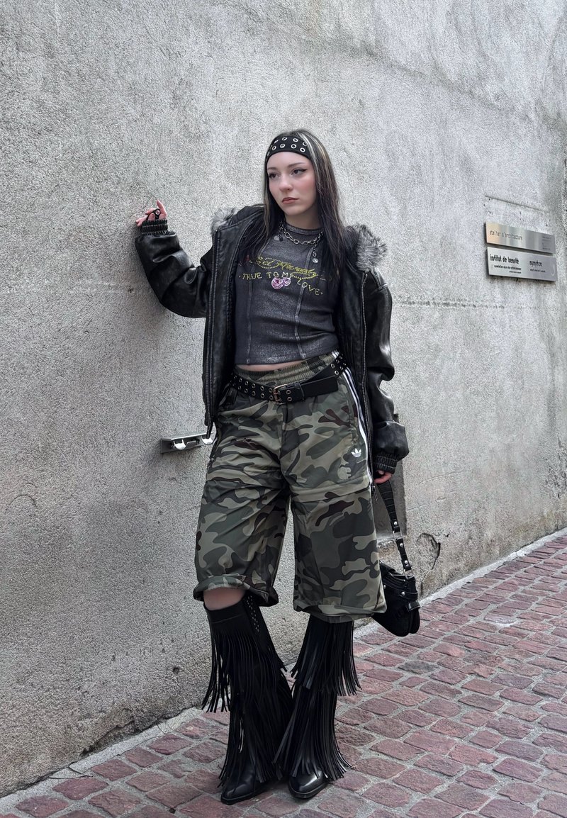 Jeune personne aux cheveux noirs et argentés appuyée contre un mur, portant une veste en cuir noire, un pantalon camouflage, des bottes à franges, et portant un petit sac noir.