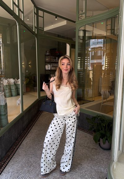 Mujer joven con cabello largo y rubio está dentro de un espacio con paredes de cristal, lleva pantalones blancos con lunares, blusa crema, sostiene un bolso negro y gafas de sol.