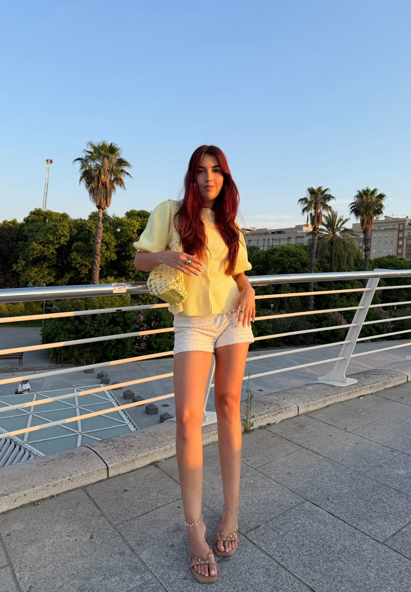 Young woman with long red hair in yellow top and white shorts standing on urban bridge railing at sunset with palm trees in background.