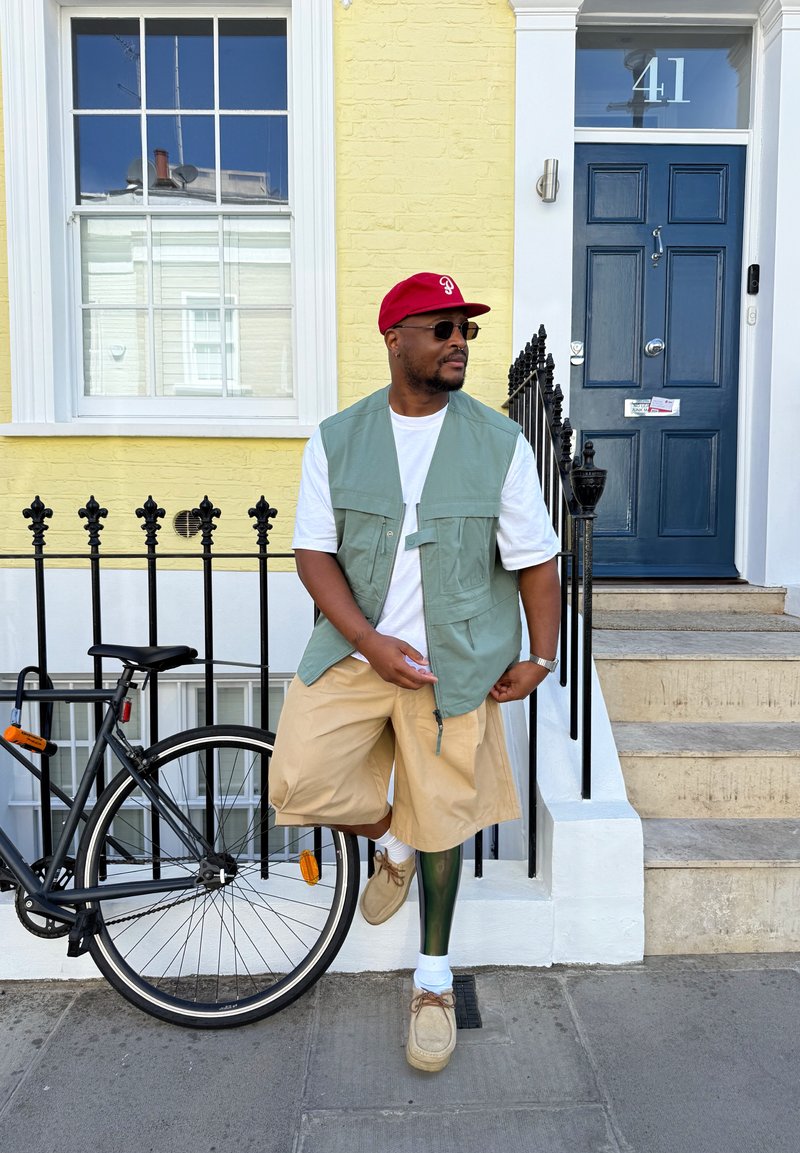 Homme portant des lunettes de soleil, une casquette rouge, un gilet vert et un short beige, debout à côté d'un vélo noir devant un bâtiment jaune avec une porte bleue numéro 41.