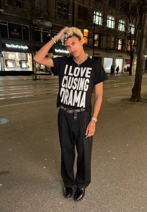 Camiseta negra con un texto blanco en negrita que dice "ME ENCANTA CAUSAR DRAMA", combinada con unos pantalones negros holgados y zapatos negros brillantes. Incluye accesorios plateados.