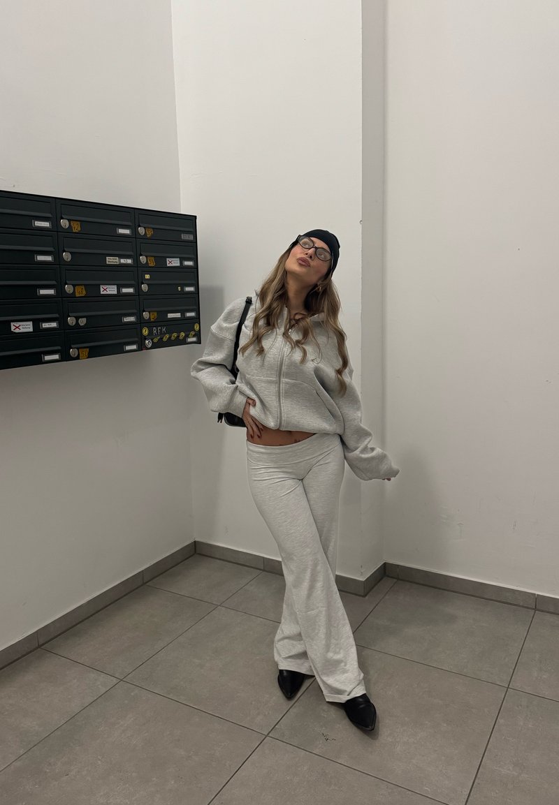 Light gray sweatshirt and matching sweatpants, soft texture, relaxed fit, worn with a black beanie and glasses, in a minimalist hallway.