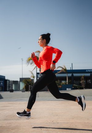 Corridore in una maglietta a maniche lunghe rossa brillante, leggings neri e scarpe da corsa nere, in movimento su un pavimento con cielo sereno e palme.