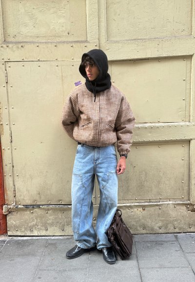 Joven con capucha negra, chaqueta marrón a cuadros, vaqueros sueltos azul claro y zapatos negros, de pie frente a una gran puerta metálica beige con un bolso marrón.