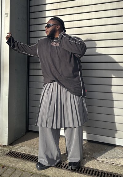 Camisa negra de manga larga de gran tamaño con costuras blancas, combinada con una falda gris plisada y pantalones de pierna ancha. Gafas de sol y zapatos negros.