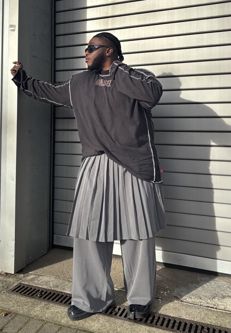 Übergroßes schwarzes Langarmhemd mit weißen Nähten, kombiniert mit einem grauen Faltenrock und weit geschnittenen Hosen. Schwarze Sonnenbrille und Schuhe.