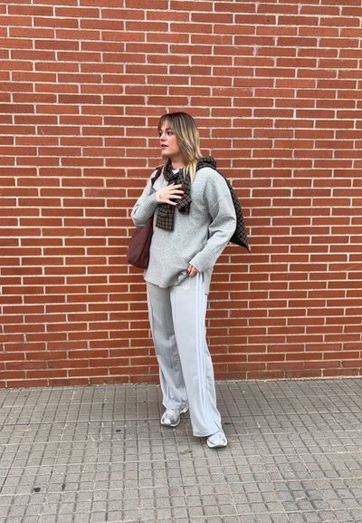 Mujer con suéter gris y pantalones de chándal blancos de Adidas está de pie sobre un pavimento de baldosas frente a una pared de ladrillo rojo, sosteniendo una bolsa marrón y una bufanda de cuadros.