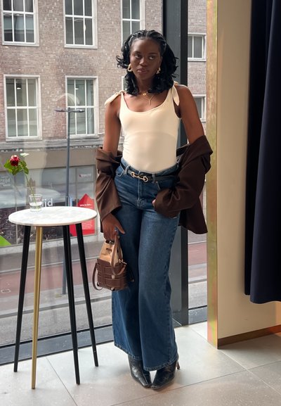 Top blanco sin mangas con tirantes delgados, chaqueta marrón de gran tamaño, jeans azules de pierna ancha, botas negras de tacón y un pequeño bolso marrón.