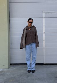 Camisa marrón de gran tamaño con botones, capucha y cuello, combinada con unos jeans de color azul claro y zapatos negros. El modelo lleva un bolso gris.