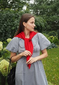 Schwarz-weißer kariertem Kleid mit Puffärmeln, ergänzt durch einen roten Schal, posiert vor einem grünen Gartenhintergrund. Schwarze Handtasche inklusive.