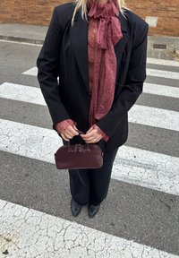 Άτομο που φοράει μαύρο σακάκι και παντελόνι, μπορντό δαντελένιο πουκάμισο και κασκόλ, κρατώντας μια μικρή μπορντό τσάντα, στέκεται σε διάβαση πεζών.