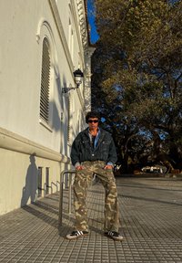 Chaqueta de mezclilla, camisa azul, pantalones de camuflaje y zapatillas negras con rayas blancas; sentado cerca de un pavimento texturizado y un edificio blanco.