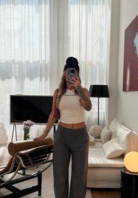 Biały prążkowany krótki top z napisem, szare szerokie spodnie w paski, czapka beanie w kolorze czarnym oraz bordowa torba. Wnętrze z sofą i dekoracyjną lampą.