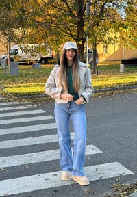 Jovem mulher com longos cabelos, vestindo um boné, uma jaqueta leve, um hoodie verde e jeans, em pé numa passadeira segurando um copo de café para levar.