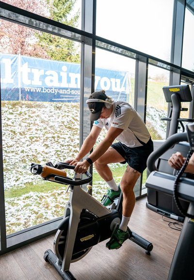 Bicicleta estática con un marco plateado y acentos en negro y naranja. Un usuario está pedaleando, vistiendo zapatos verdes, una camiseta blanca y auriculares.