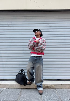 Persona con chaqueta de cuadros oversized, jeans holgados, gorra y gafas de sol, de pie frente a una persiana cerrada con una bolsa negra en el suelo.