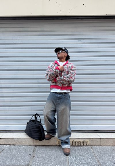 Persona con chaqueta de cuadros oversized, jeans holgados, gorra y gafas de sol, de pie frente a una persiana cerrada con una bolsa negra en el suelo.