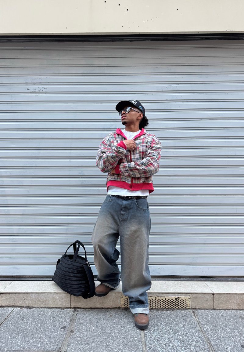 Personne portant une veste surdimensionnée à carreaux, un jean ample, une casquette et des lunettes de soleil, se tenant contre un volet fermé avec un sac noir posé au sol.