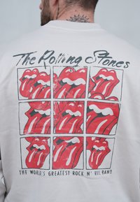 Sudadera blanca con un gráfico de nueve logotipos de lengua roja, con la inscripción "The Rolling Stones" arriba y "LA MEJOR BANDA DE ROCK N' ROLL DEL MUNDO" abajo.