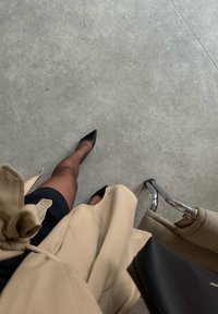 Personne portant des talons noirs pointus, des collants noirs transparents, une jupe noire et un trench-coat beige, debout sur un sol gris à côté d'une chaise beige.
