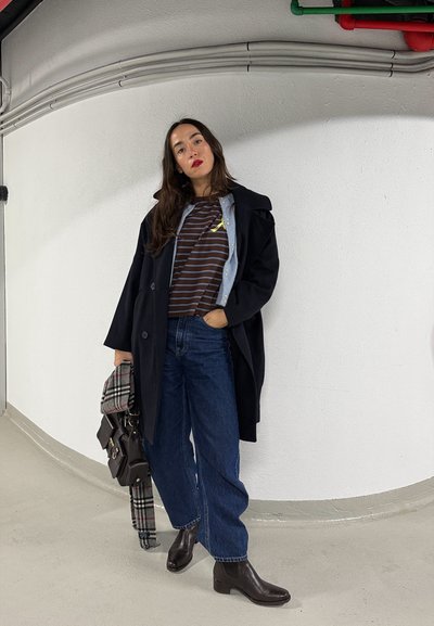 Mujer de cabello largo y oscuro, vestida con abrigo negro, camiseta de rayas, jeans azules anchos y botines oscuros, sosteniendo una bolsa y una bufanda a cuadros, de pie en el interior.