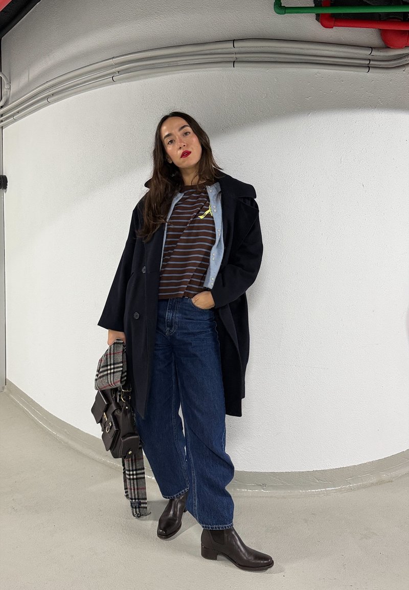 Vrouw met lang donker haar, gekleed in een zwarte jas, gestreept shirt, wijde blauwe jeans en donkere enkellaarzen, die een tas en een geruit sjaal vasthoudt, staand binnen.