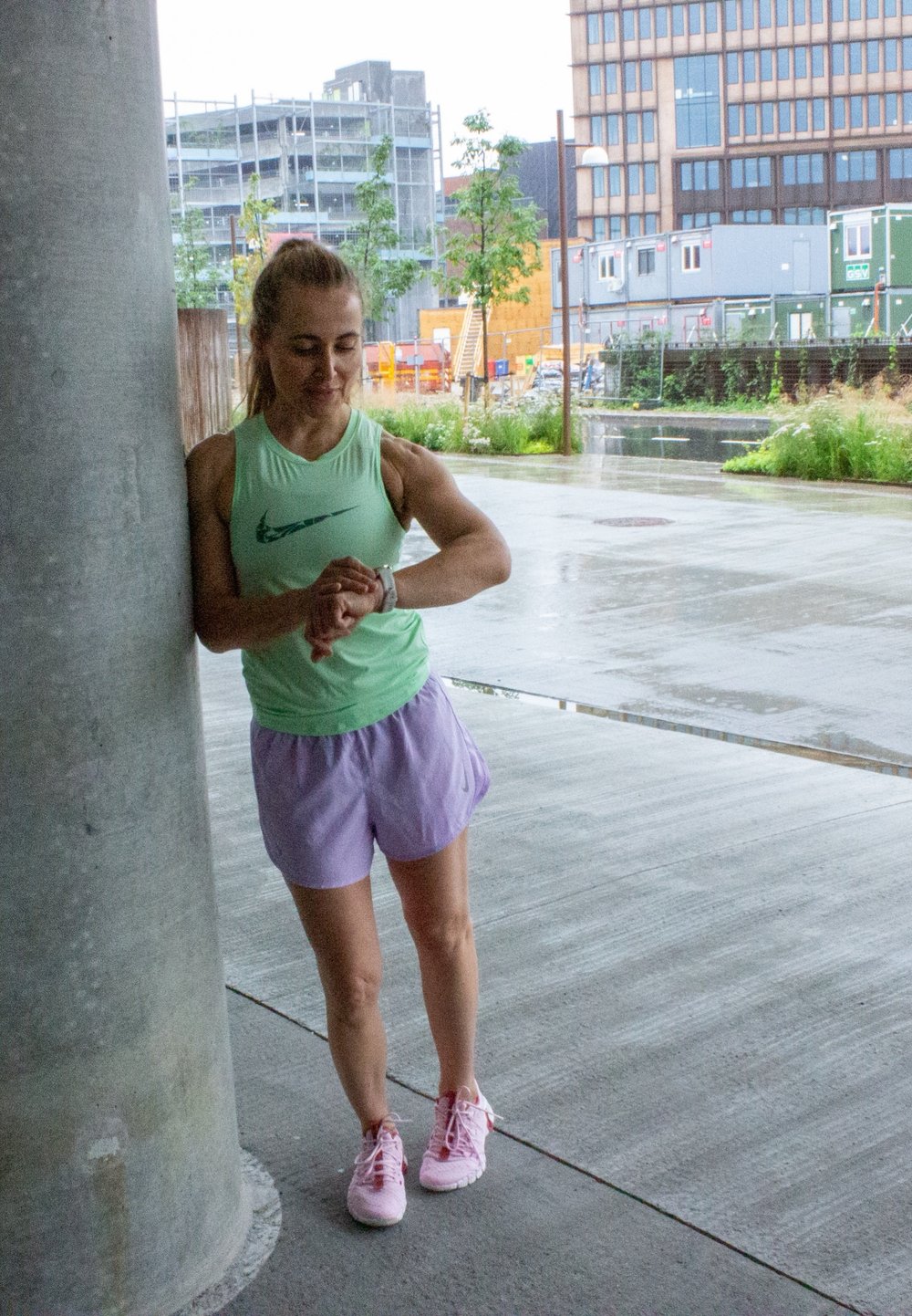 Atleta vestida com uma blusa sem mangas verde-clara e calções lavanda, a ver as horas num relógio. Ela usa sapatilhas de corrida rosa, encostando-se a um pilar de betão.