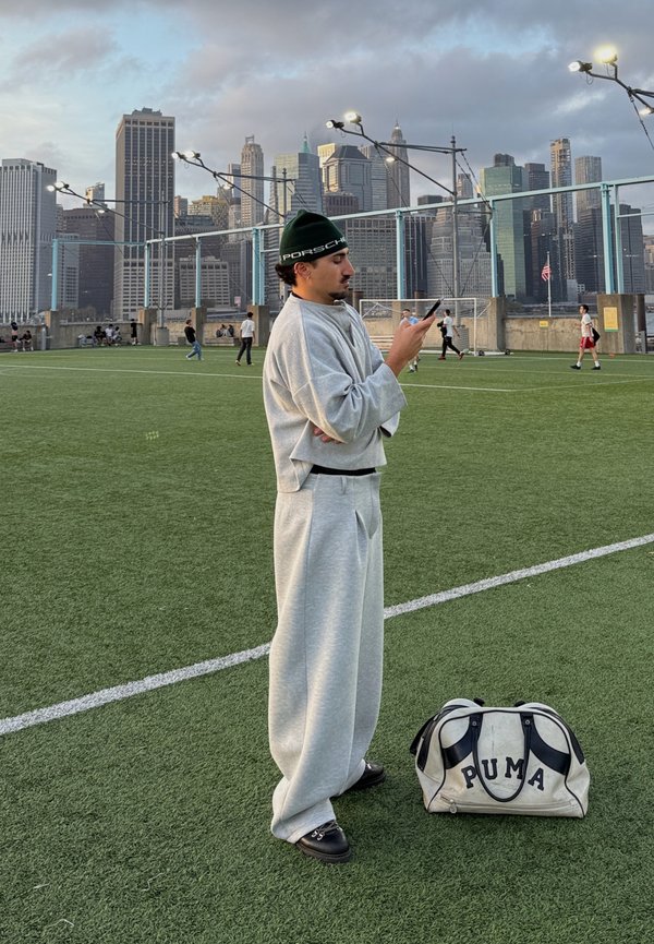 Light grey sweatshirt and trousers worn with black shoes. A black and white Puma holdall rests on the ground. Urban skyline in the background.
