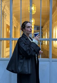 Trench-coat noir au design structuré, associé à un pull léger. Tenant une tasse bleue, l'arrière-plan présente une architecture ornée et un éclairage chaleureux.