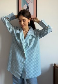 Ανοιχτό μπλε και λευκή ριγέ φανέλα σε υπερμεγέθη μέγεθος, με μακριά μανίκια και κουμπωτές μανσέτες, τσέπη στο στήθος, φορεμένη με μπλε τζιν. Μινιμαλιστικός σχεδιασμός.