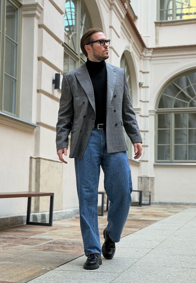 Homme portant un blazer gris à carreaux, un col roulé noir, un jean bleu et des chaussures noires, marchant en plein air dans une cour avec des fenêtres en arc.