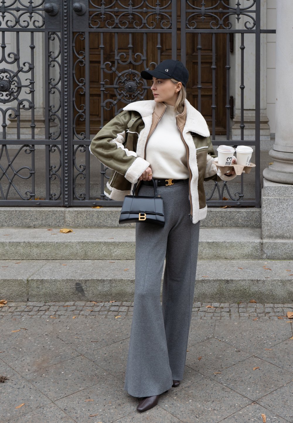 Casaco de shearling verde com forro branco, suéter beige de gola alta, calças largas cinzas, bolsa preta, segurando copos de café, em pé à frente de um portão ornamentado.