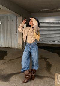 Beige bomberjakke over en tettsittende beige topp, kombinert med løse blå denimjeans og brune hælboots. En gul veske fullfører antrekket.
