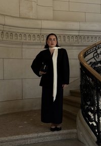 Blazer nero su misura e gonna lunga con una sciarpa bianca, in piedi su scale di marmo con una ringhiera decorativa, mentre tiene una piccola borsa nera.