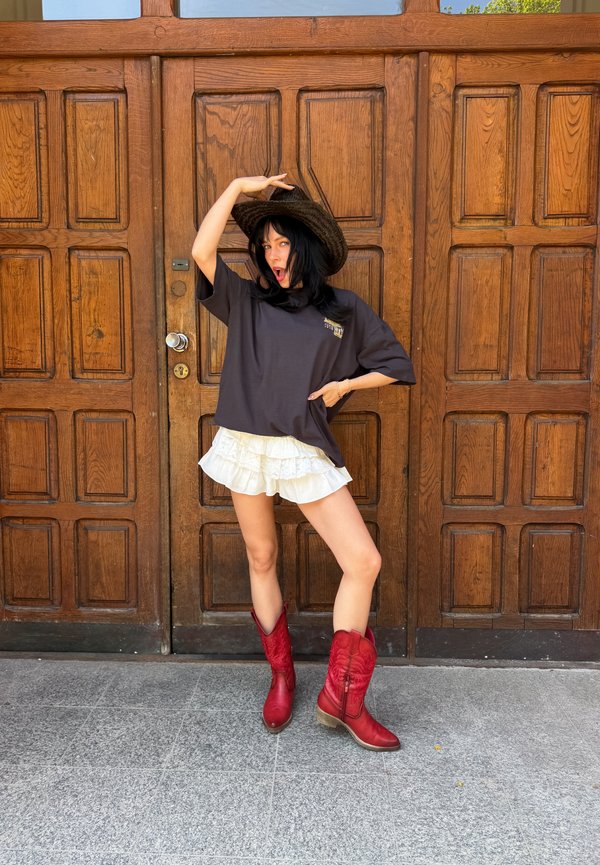 Kvinde iført sort hat, oversize mørk t-shirt, hvid flæset nederdel og røde cowboystøvler poserer foran en stor trædør.