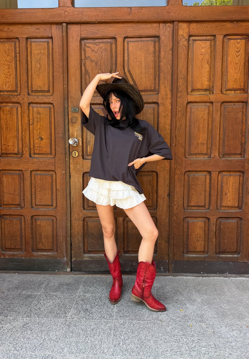 Γυναίκα που φοράει μαύρο καπέλο, φαρδιά σκούρα μπλούζα, λευκή φούστα με βολάν και κόκκινες καουμπόικες μπότες ποζάρει μπροστά από μια μεγάλη ξύλινη πόρτα.