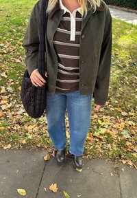 Khaki zelena jakna, rjava in bela črtasta polo majica, modri široki jeans, črna prešita torba za ramo, črne škornje, jesenski listi v ozadju.
