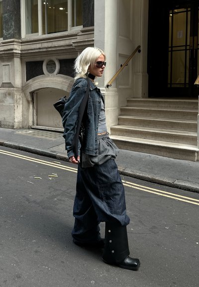 Chaqueta de denim y pantalones holgados en azul oscuro, camiseta gris y botas negras hasta la rodilla con botones plateados; combinado con gafas de sol y un bolso negro.