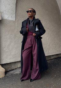 Femme portant un manteau noir surdimensionné, de grandes lunettes de soleil, un pantalon large bordeaux et un haut assorti, tenant un petit sac à main noir contre un mur texturé.